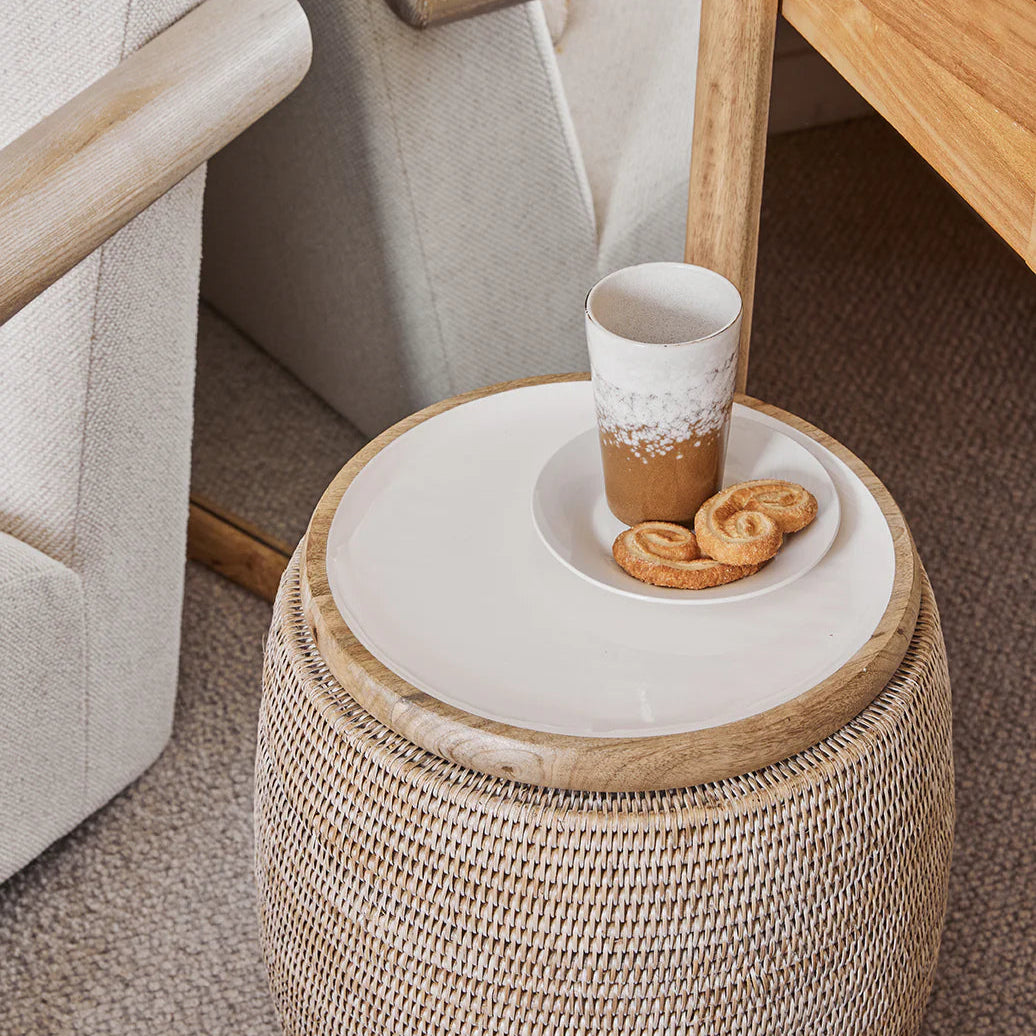 Small round woven table with a ceramic cup and cookies on a carpeted floor.
